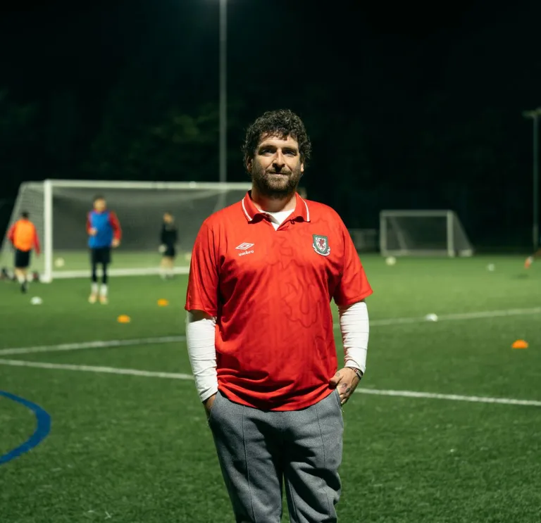 Le fondateur du club, Delwyn Derrick, debout sur un terrain de football, tandis qu'un match se déroule en arrière-plan.