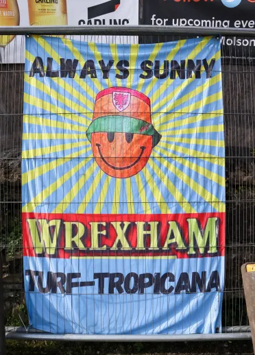 A sign showing a smiley face wearing a bucket hat and text that reads "ALWAYS SUNNY WREXHAM TURF-TROPICANA"