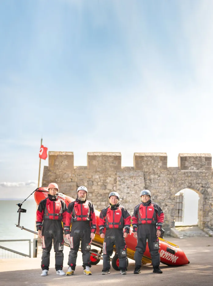 Four men dressed in sea rescue clothing stand in front of a stationary lifeboat on land in front of  a castle wall