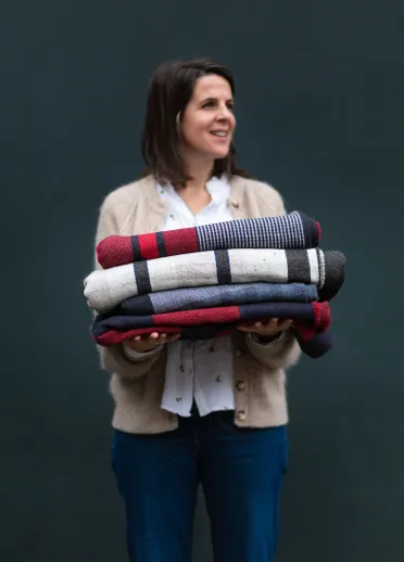 Welsh textile artist Llio James standing in front of a dark coloured wall, holding a stack of neatly folded blankets that she has made