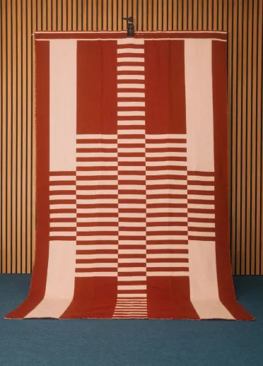 A large hand-woven red and white blanket in a striped pattern created by Welsh textile artist Llio James. The blanket is hanging on a wooden wall