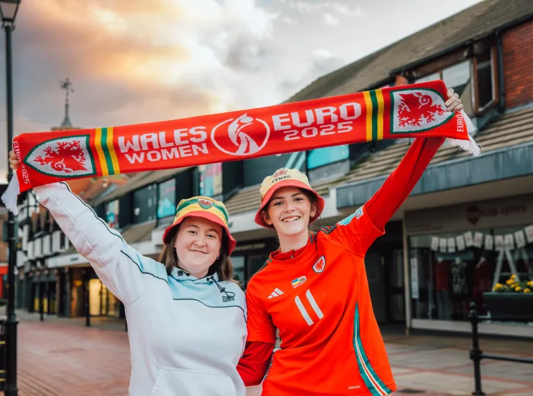 Two people holding a 'WALES WOMEN EURO 2025' scarf in a shopping area, with shops and a cloudy sky behind them.