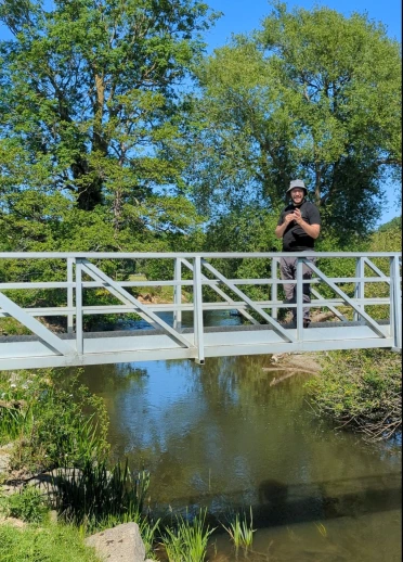 Joe standing on a white metal footbridge over a river, surrounded by trees on a sunny day.