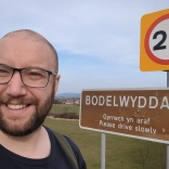 Joe taking a selfie beside a “Bodelwyddan” village sign in a grassy landscape.