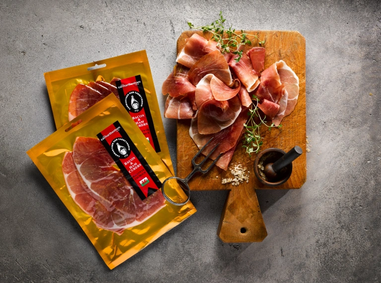 Two packets of ham and a wooden board displaying carved ham.