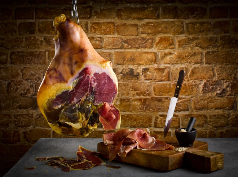 Ham hanging from a hook and a knife next to sliced ham.