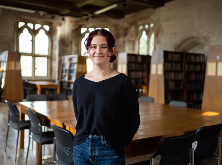 A student in a black top and blue jeans standing in front of a table inside the college.