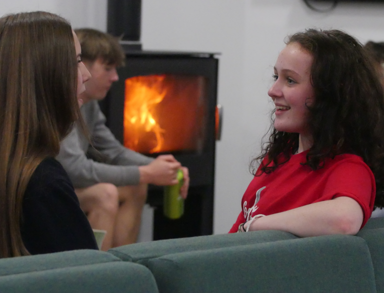 People sat on a sofa and talking with log burner in background.