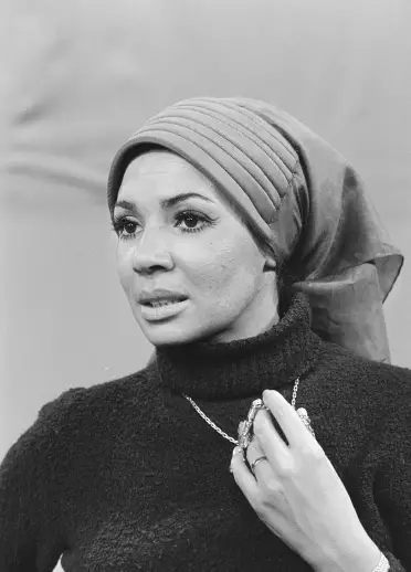 A black and white photo of Bassey wearing a scarf over her hair and with her jewelled hand holding onto her necklace.