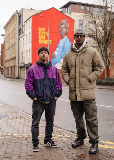 Two men standing in front of a mural of a pregnant woman wearing a blue football shirt.