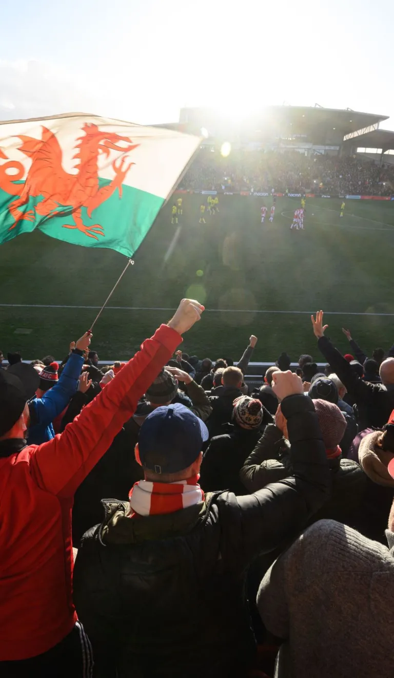 Une foule de personnes jubile pendant un match de football, avec un drapeau gallois flottant au milieu d'eux dans le stade.