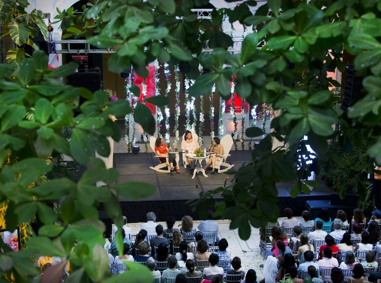 Three people sat on chairs round a table on a stage with an audience watching.