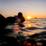 Surfer in wetsuit floating on the surface of the sea, silhouetted by the sunset on the horizon