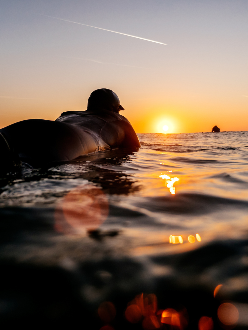 Surfer in wetsuit floating on the surface of the sea, silhouetted by the sunset on the horizon