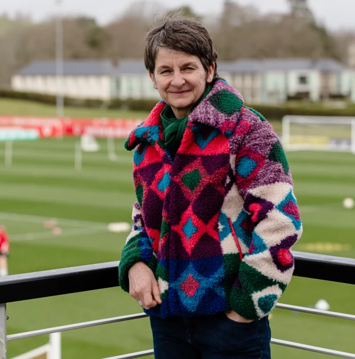 Dressed in a colourful jacket, Laura stands with her back to a football field with one hand in her pocket and smiling into the camera