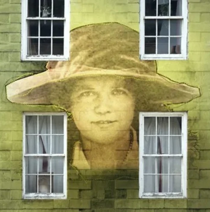 Lucy is a hologrammed image against a wall with four windows, she is wearing a wide brimmed hat and staring directly into the camera 