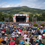 A crowd of people sat in a field looking towards an outdoor festival stage with a hill in the background
