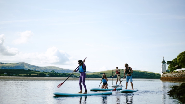 Young people standing on paddleboards in the water surrounded by soft hills and a tower. 