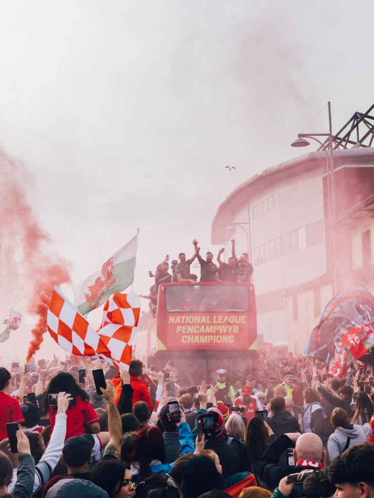 A tour bus with a sports team driving past a stadium, through throngs of fans waving flags and letting off flares