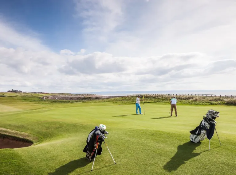 An outdoor scene of a men playing golf set against a blue sky with the sea in the background 