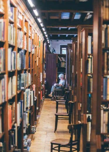 A lady sitting and reading in a library, surrounded by high shelves full of books.