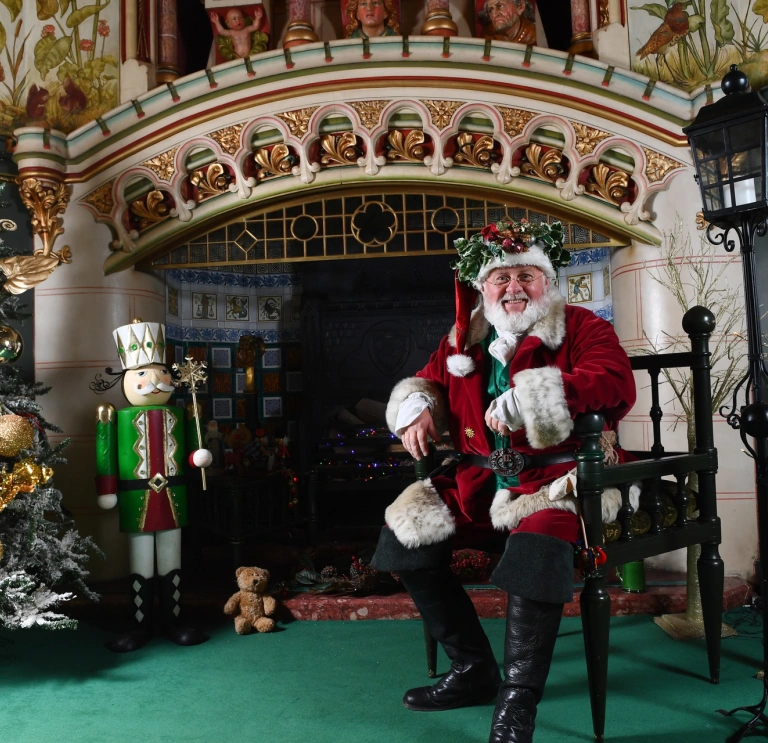 Siôn Corn sitting on a chair by a fire and Christmas decorations.