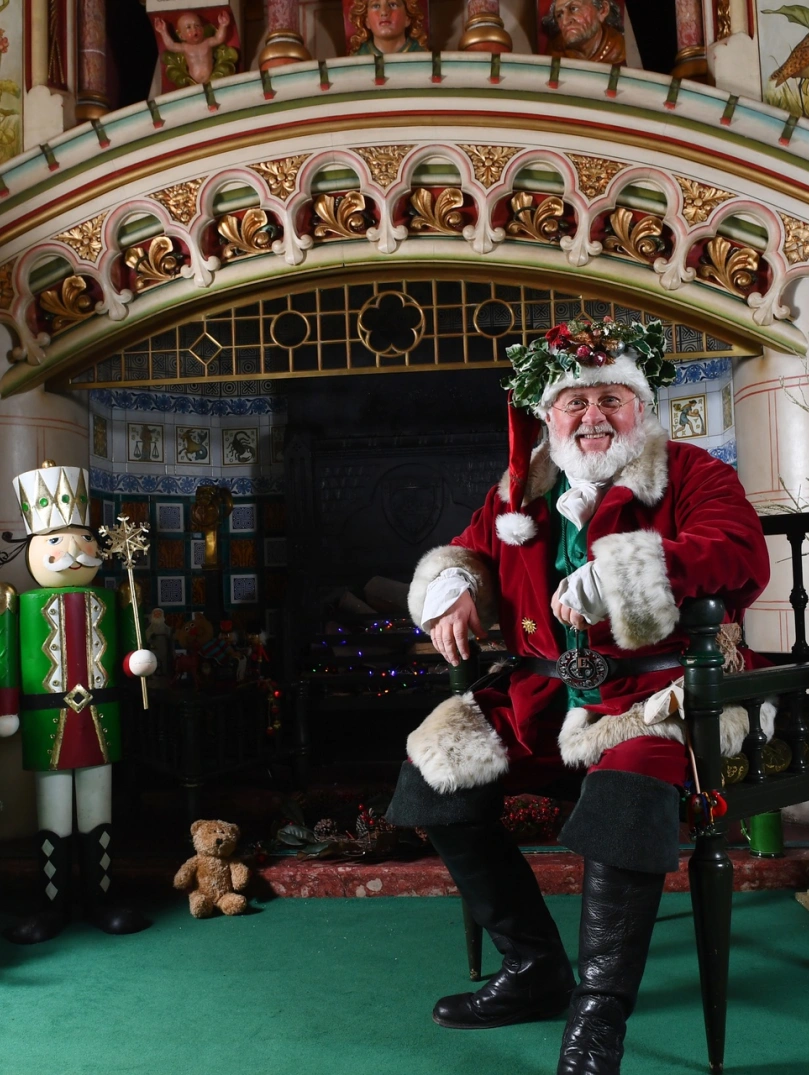 Siôn Corn sitting on a chair by a fire and Christmas decorations.