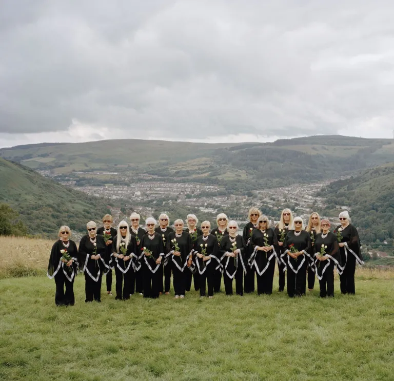 The Dare to Sing women's choir dressed in black, standing on a patch of green in front of rolling Welsh valleys