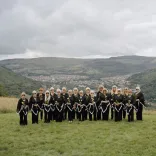 The Dare to Sing women's choir dressed in black, standing on a patch of green in front of rolling Welsh valleys