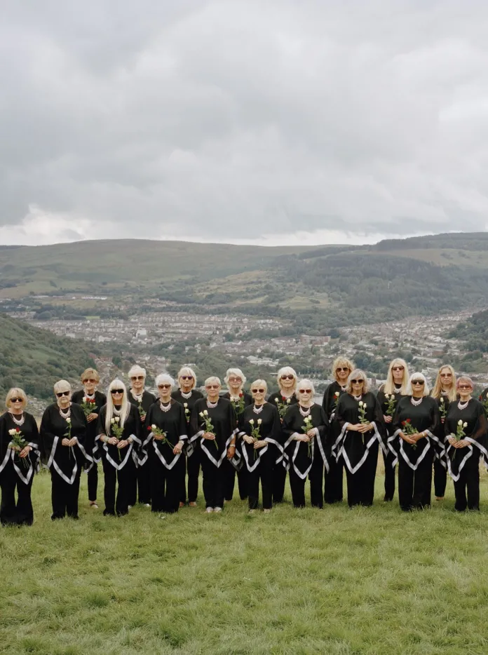 The Dare to Sing women's choir dressed in black, standing on a patch of green in front of rolling Welsh valleys