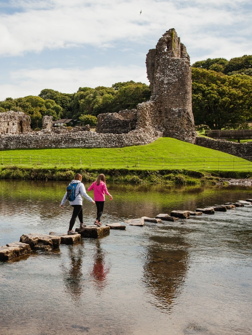 Horses and walkers in front of a ruined castle surrounded by green fields and water.