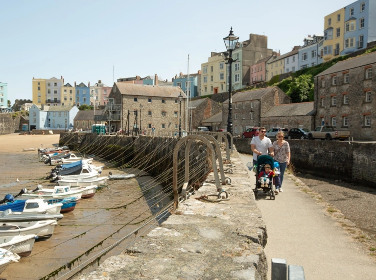 A family walking with a baby in a pushchair along a beachside with boats moored up on the sand and colourful houses in the background