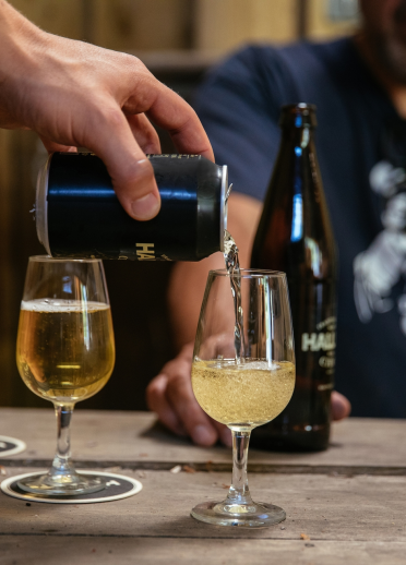 A hand pouring a can of Hallets Cider into a glass on a wooden table.