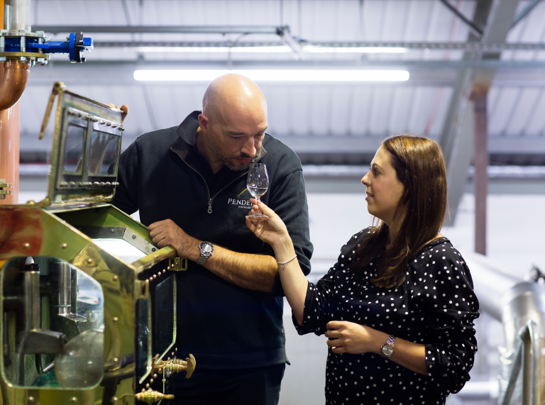 man leaning on machinery smelling whisky as woman holds glass for him 