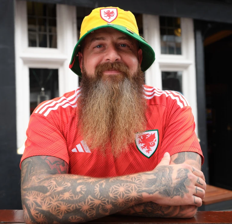 A man with a long beard and tattooed arms smiling at the camera, while wearing a Welsh bucket hat and Wales football strip