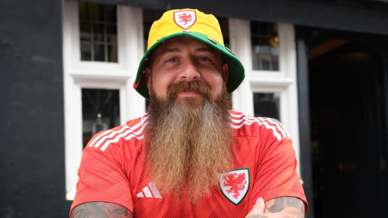 A man with a long beard and tattooed arms smiling at the camera, while wearing a Welsh bucket hat and Wales football strip