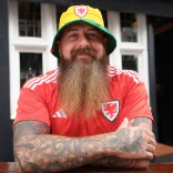 A man with a long beard and tattooed arms smiling at the camera, while wearing a Welsh bucket hat and Wales football strip