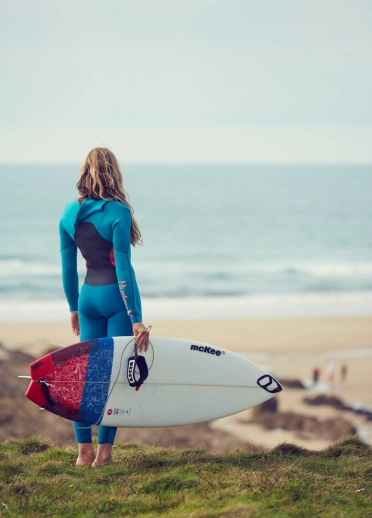 A person in a wetsuit holding a surfboard looks out over a beach and a blue sea in the distance