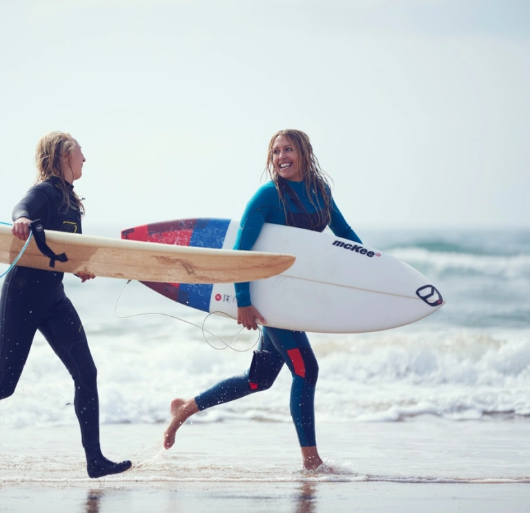 Two people wearing wetsuits and holding surfboards run through water across the sand on a beach
