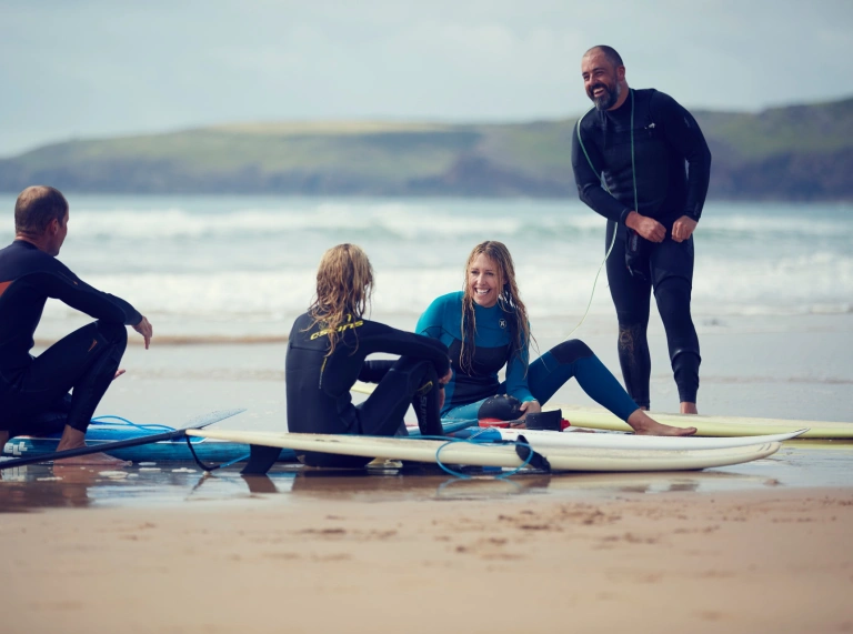Four people wearing wetsuits and holding surfboards sitting on a beach, having a nice time