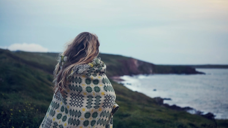 A person wrapped in a Welsh blanket, looking to a far out sea