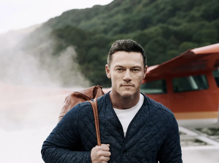 Luke Evans walking out of small aircraft with smoke behind him